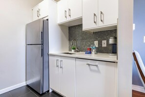 A kitchen with white cabinets, a stainless steel refrigerator, a tiled backsplash, and a countertop with a sink and various items. - Landing | Incredible 1BD, Gym (New Haven)