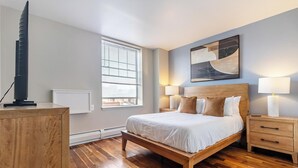 Ein modernes Schlafzimmer mit einem Holzbett, einem Fernseher auf einem Standfuß, einem Fenster mit Jalousien und einem Gemälde an der Wand.