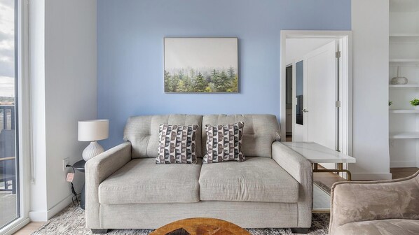 Un salon avec un canapé gris, des coussins à motifs, un tableau encadré au mur et une fenêtre donnant sur un paysage.