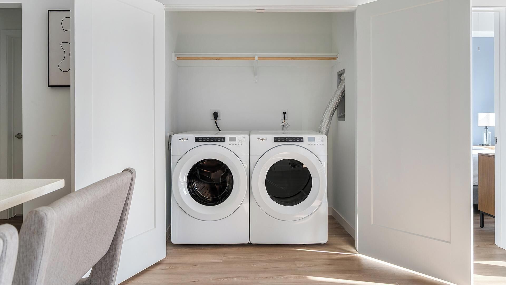 A compact laundry room with two front-loading Whirlpool washers, a wooden shelf above, and a chair to the left.
