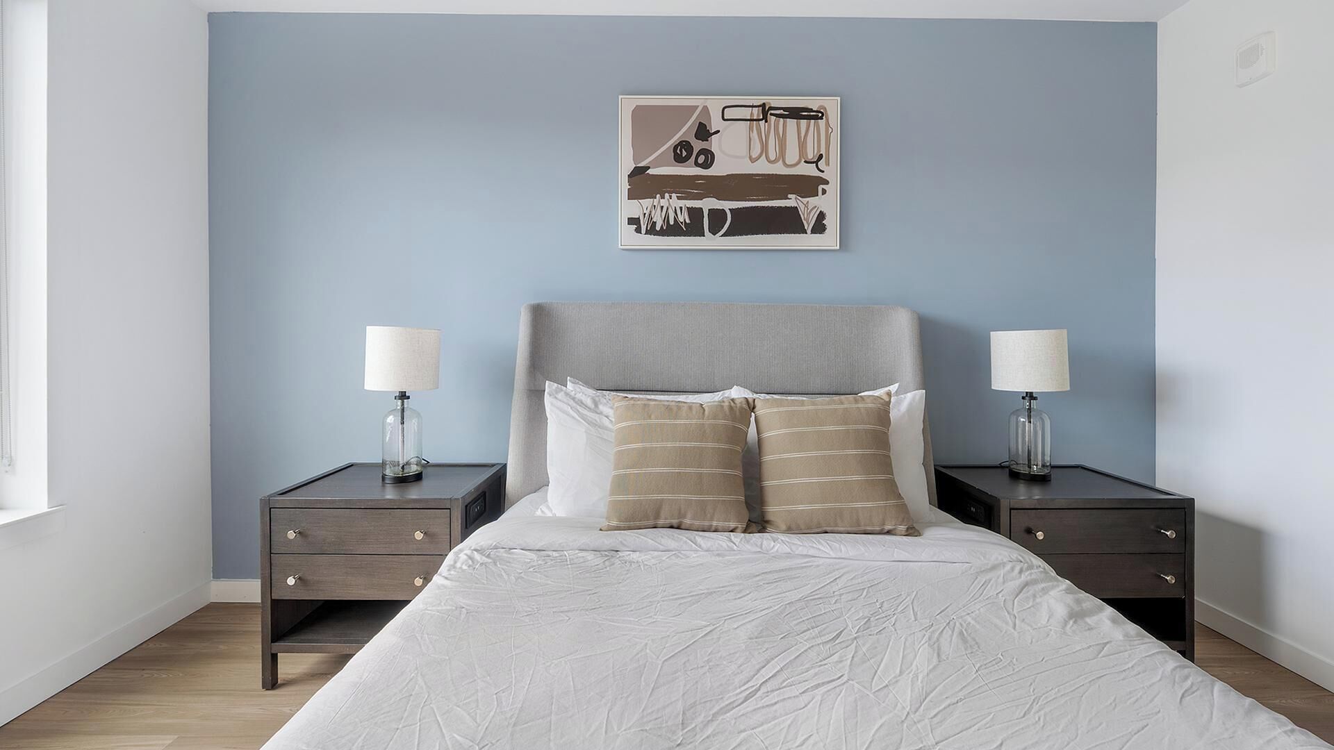 Een slaapkamer met een bed, twee nachtkastjes, twee lampen, een hoofdbord en een kunstwerk aan de muur.