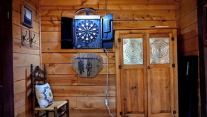 Interior de una cabaña de madera con dardos, un sillón con cojín estampado y una puerta con paneles de vidrio decorativos.
