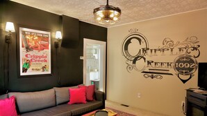 Una sala de estar con un sofá gris, cojines rojos, una bandeja con un objeto negro, y una pared con un cartel que dice 'Cripple Creek' y el logotipo de 'Ripples'.