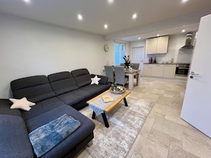 A modern living room with a dark grey sofa, a wooden coffee table, and a dining area with a small table and chairs. A kitchen with cabinets and appliances is visible in the background. - Charming apartment on the hill in one of the village's oldest streets (Bad Bentheim)