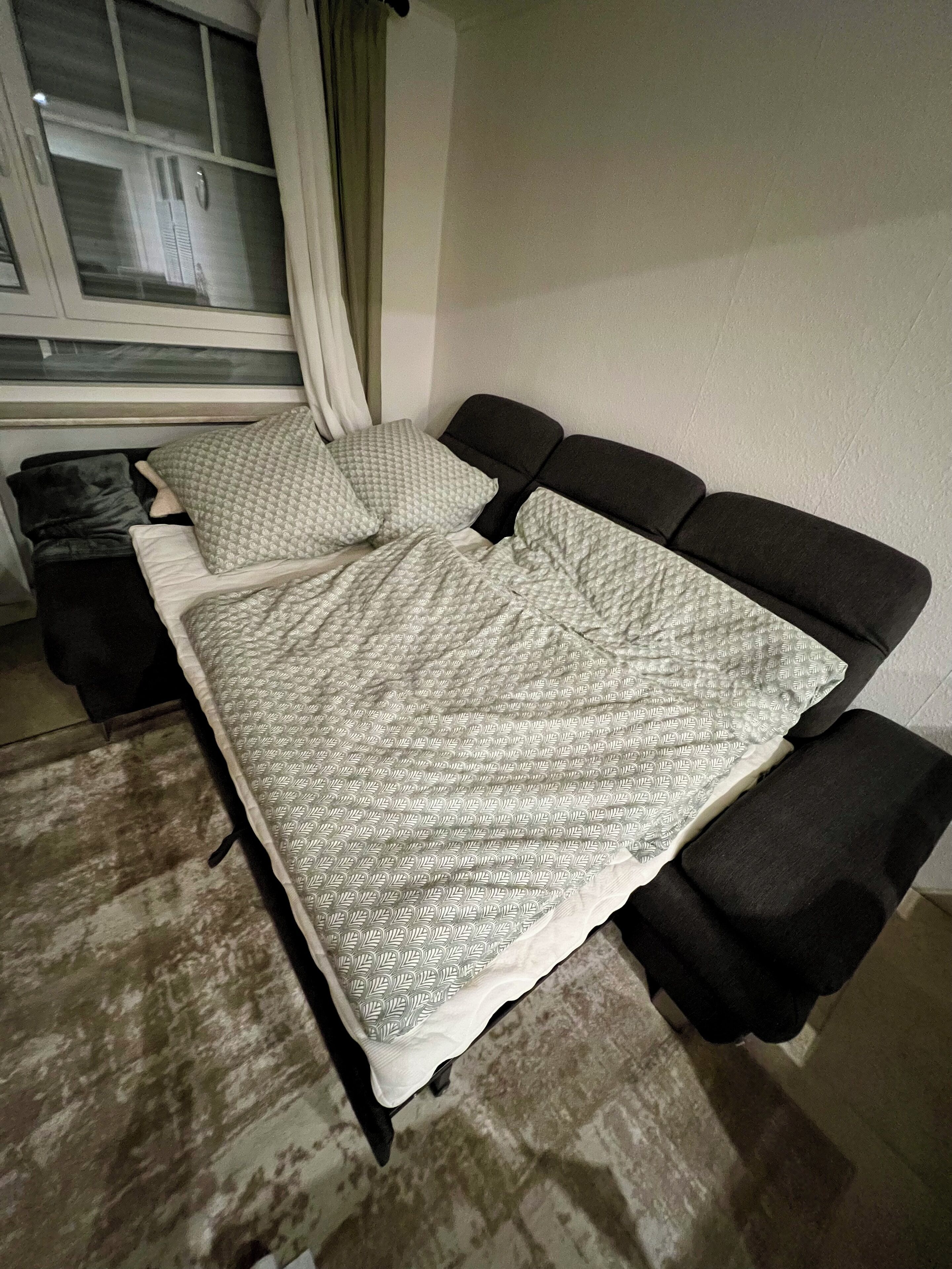 Ein Bett mit einem gemusterten Überwurf und Kissen, ein Fenster mit Vorhängen und ein Blick nach draußen.