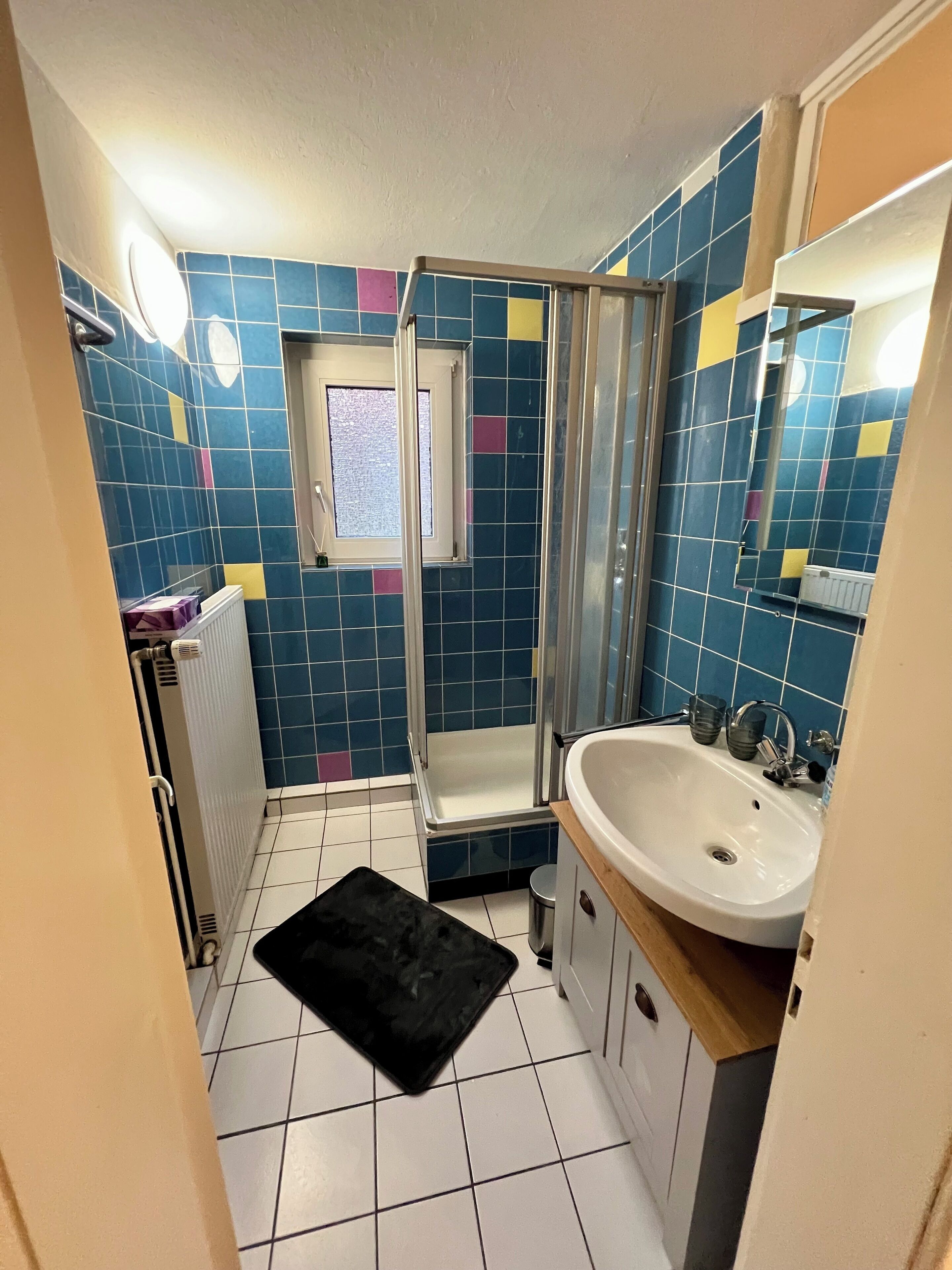 Een badkamer met blauwe en gele tegels, een witte wastafel, een douchecabine en een klein raam.