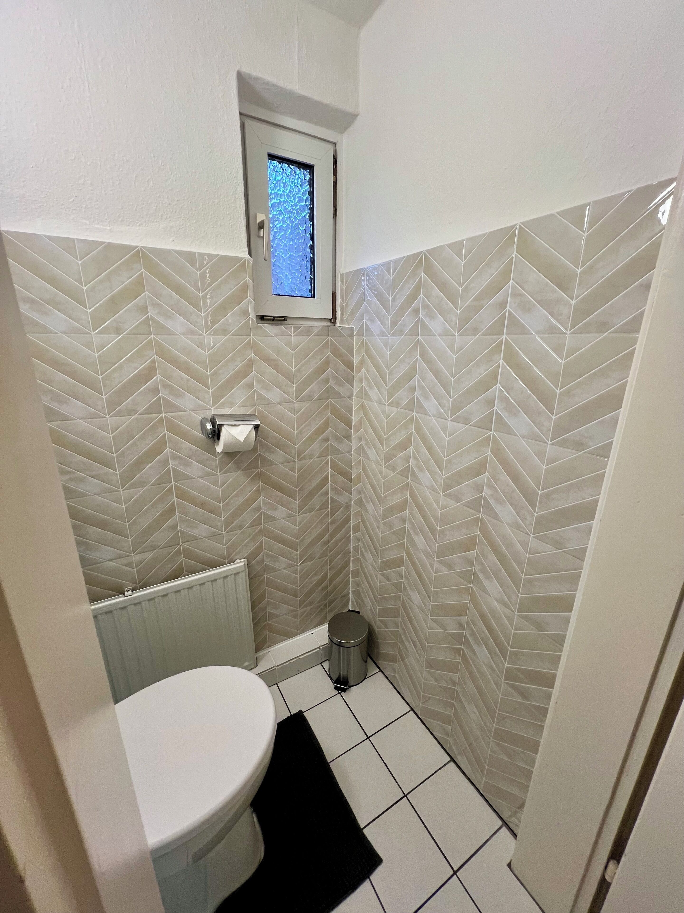 Ein Badezimmer mit einer weißen Toilette, einem kleinen Fenster und einer Wand mit Fischgrätmuster-Fliesen.