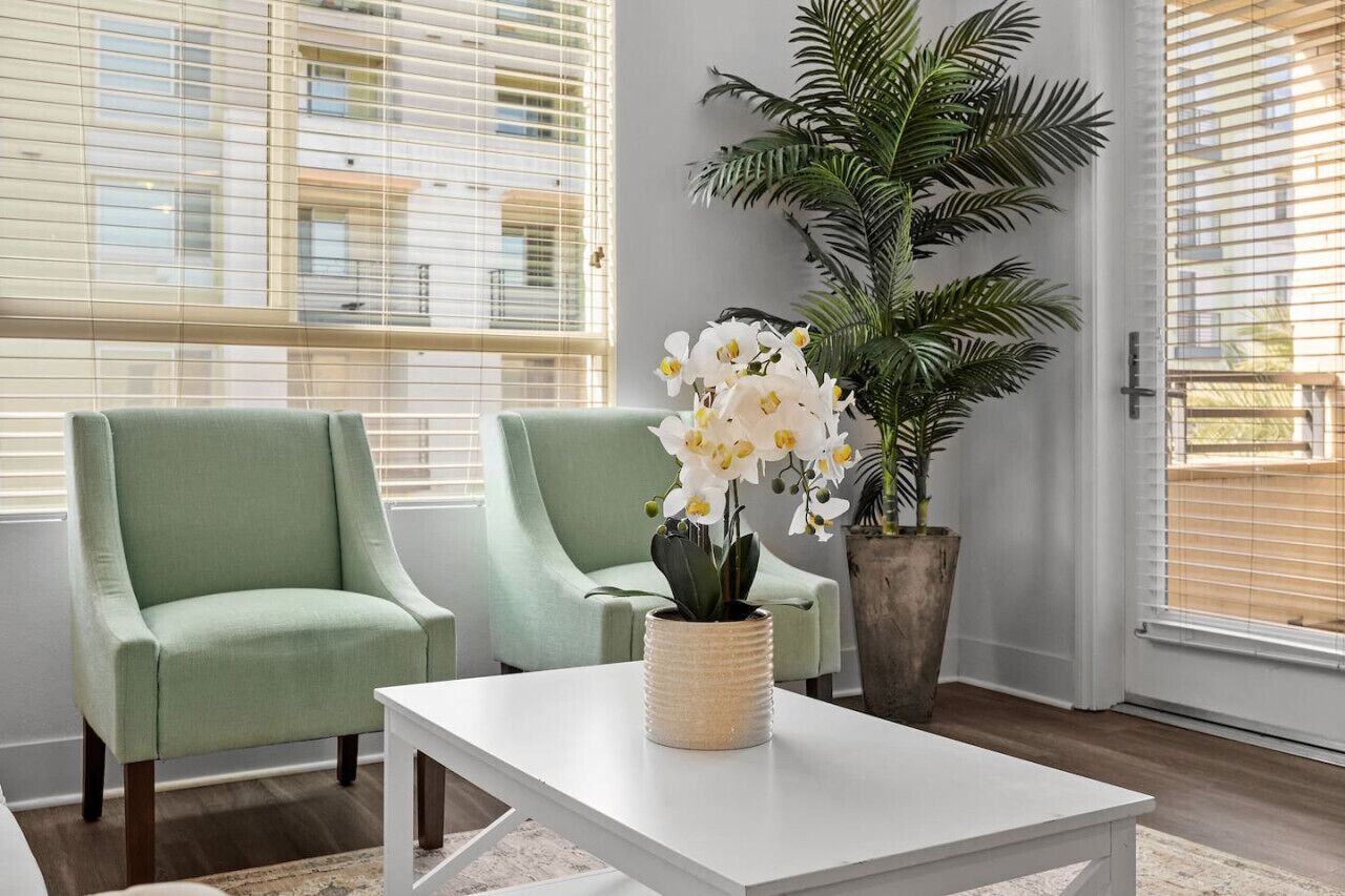 Une pièce avec deux fauteuils verts, une table basse blanche et une grande plante en pot ornée de fleurs blanches.