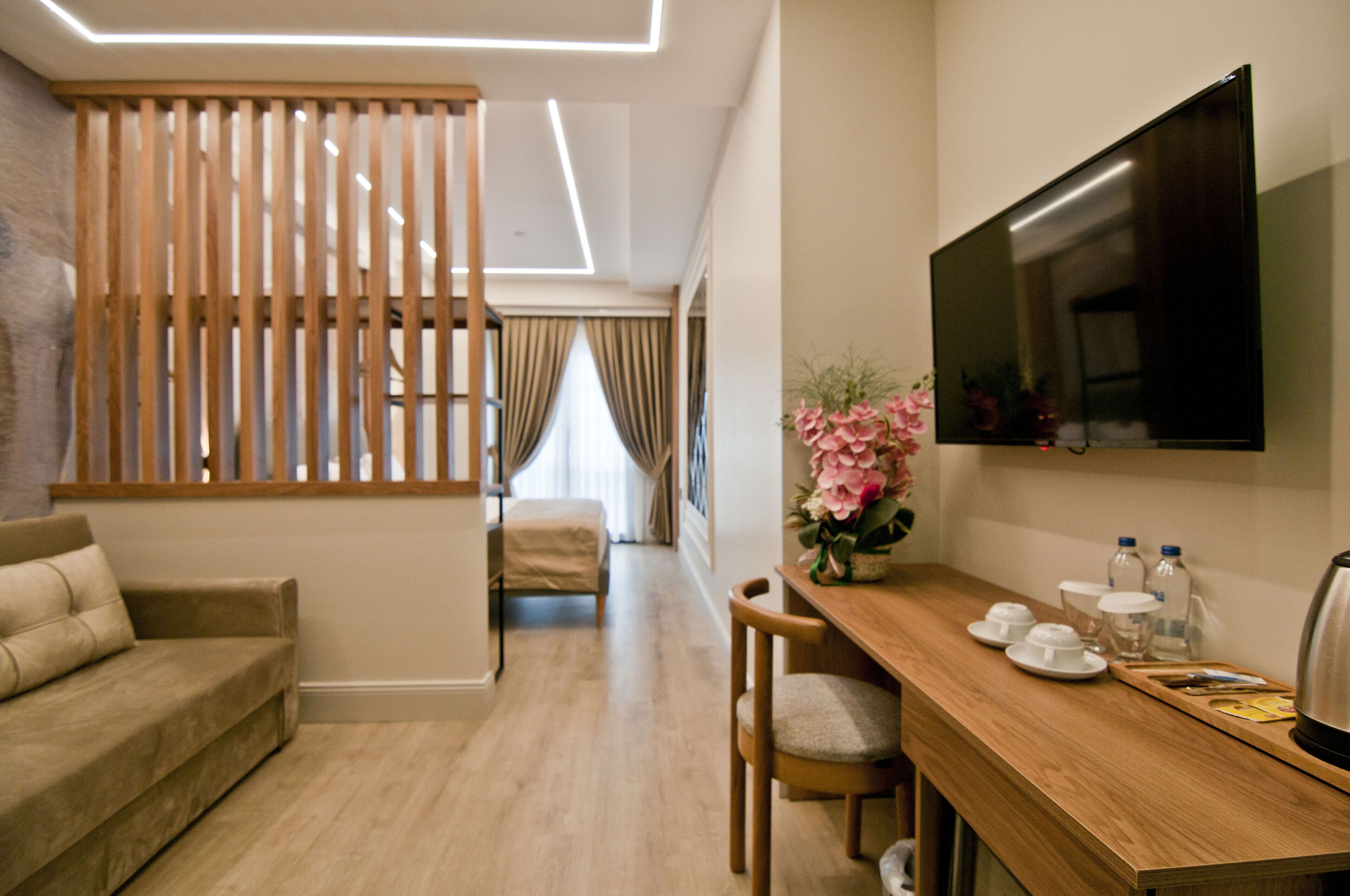 A modern hotel room with a wooden partition, a sofa, a dining table with a TV, and a vase of flowers.