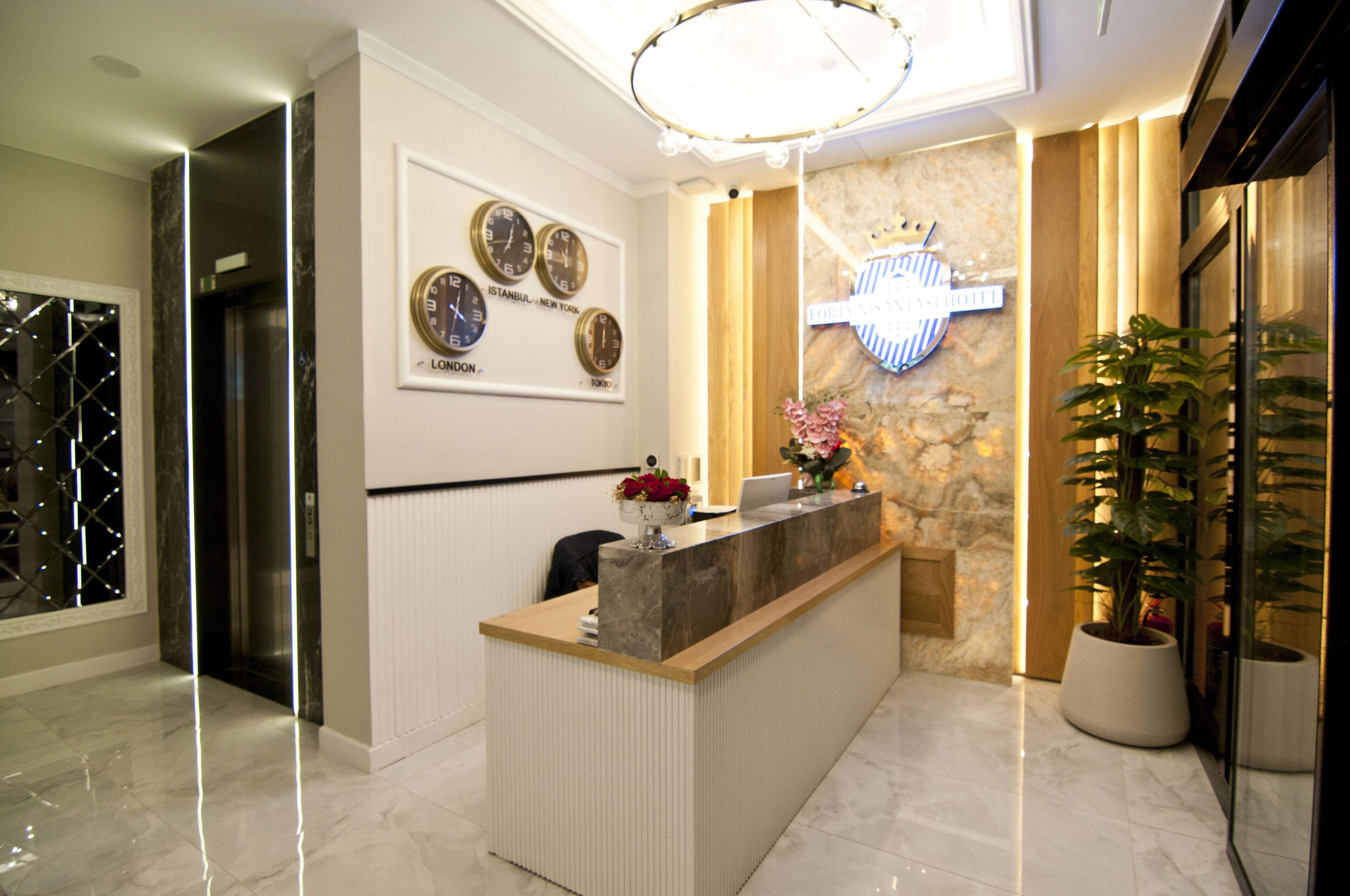 A hotel reception area with a marble reception desk, a wall clock displaying multiple time zones, a crown emblem, and a potted plant.