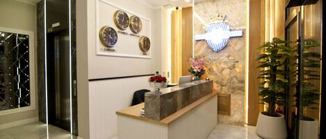 A hotel reception area with a marble reception desk, a wall clock displaying multiple time zones, a crown emblem, and a potted plant.