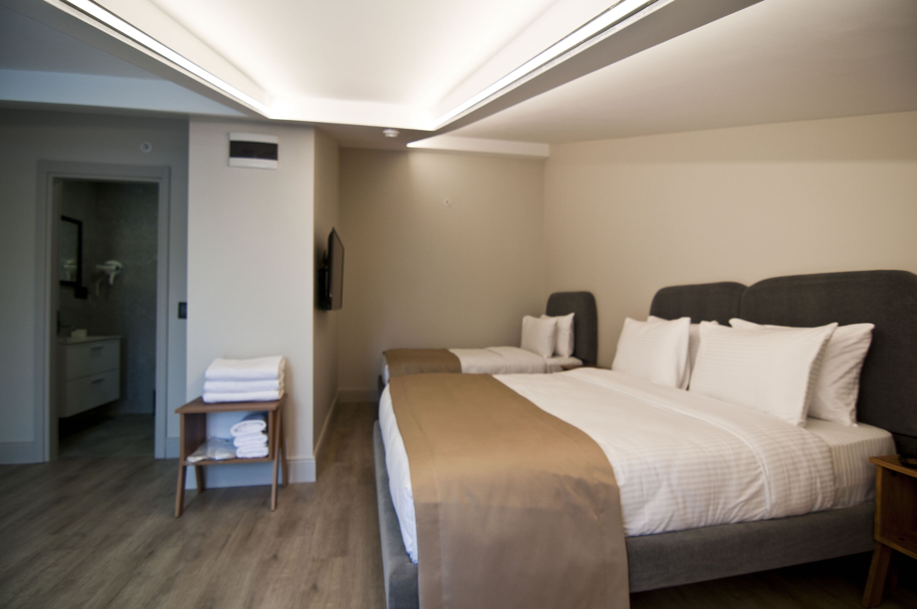 A modern hotel room with a large bed, a bedside table, a small shelf with towels, and a bathroom visible through an open door.