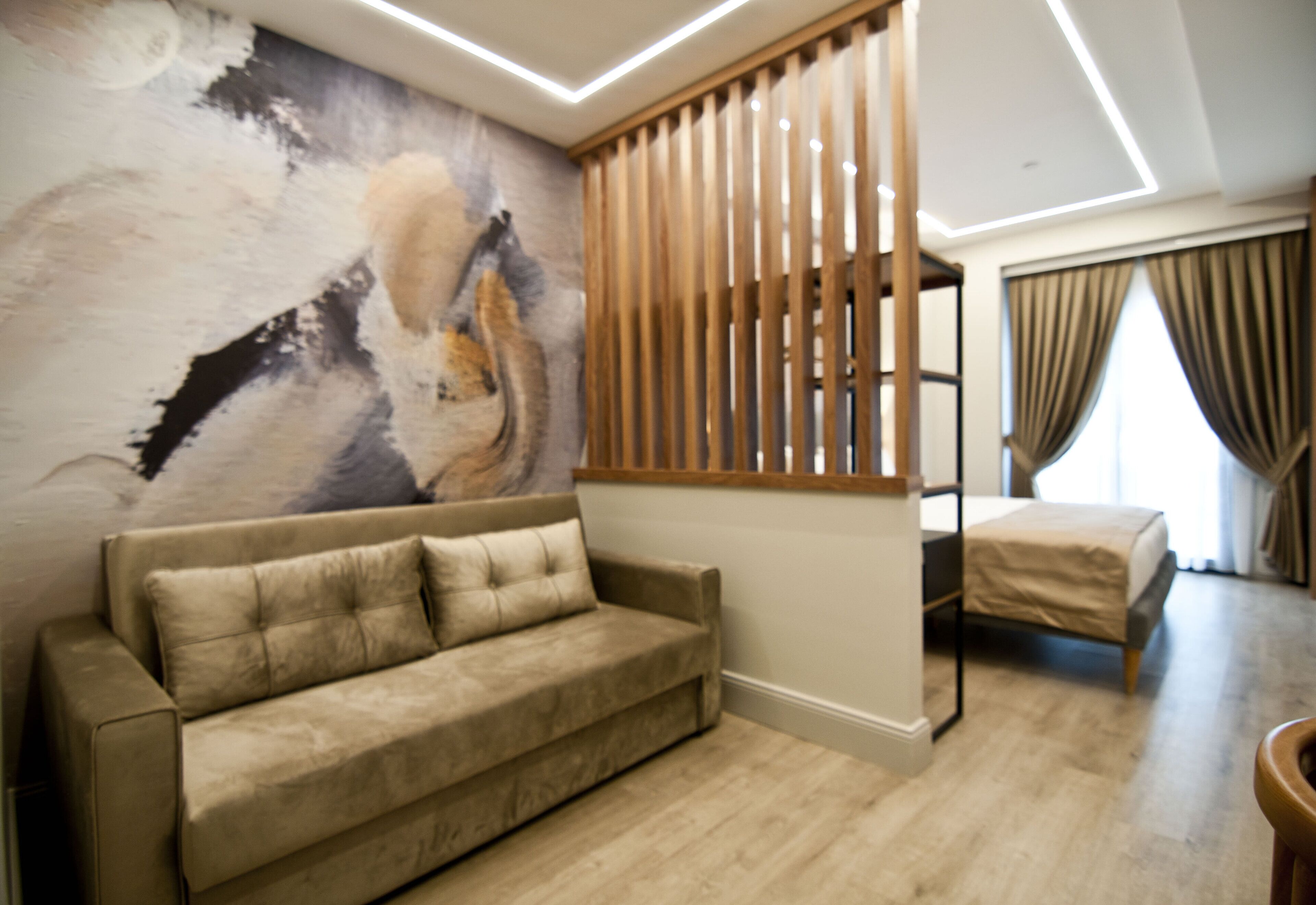 Quarto moderno com sofá bege, divisória de ripas de madeira, cama com cobertura bege e uma grande pintura abstrata na parede.
