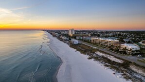 A coastal hotel with a beachfront view, a road parallel to the beach, and a large building in the distance. - H2O Living•Pool & Hot Tub•Game Room•Private Beach •Free Chair Service+Beach Gear (Miramar Beach)
