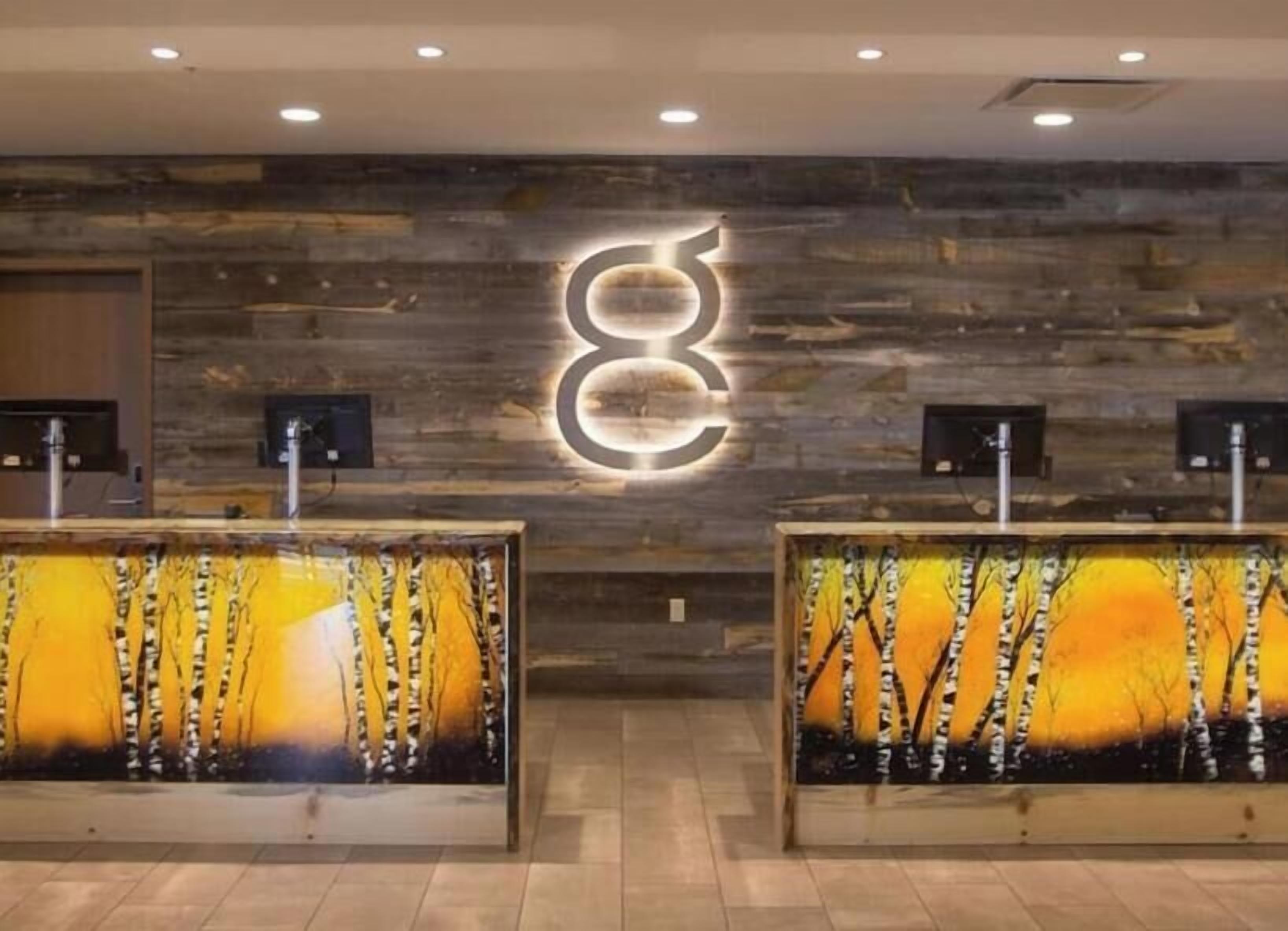 A reception area with a wooden wall and a large illuminated 'EC' sign. There are two reception desks with framed artwork depicting birch trees and a sunset.