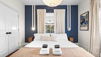 A bedroom with a large bed, two bedside tables, a window with curtains, a wall-mounted light fixture, and a framed picture.