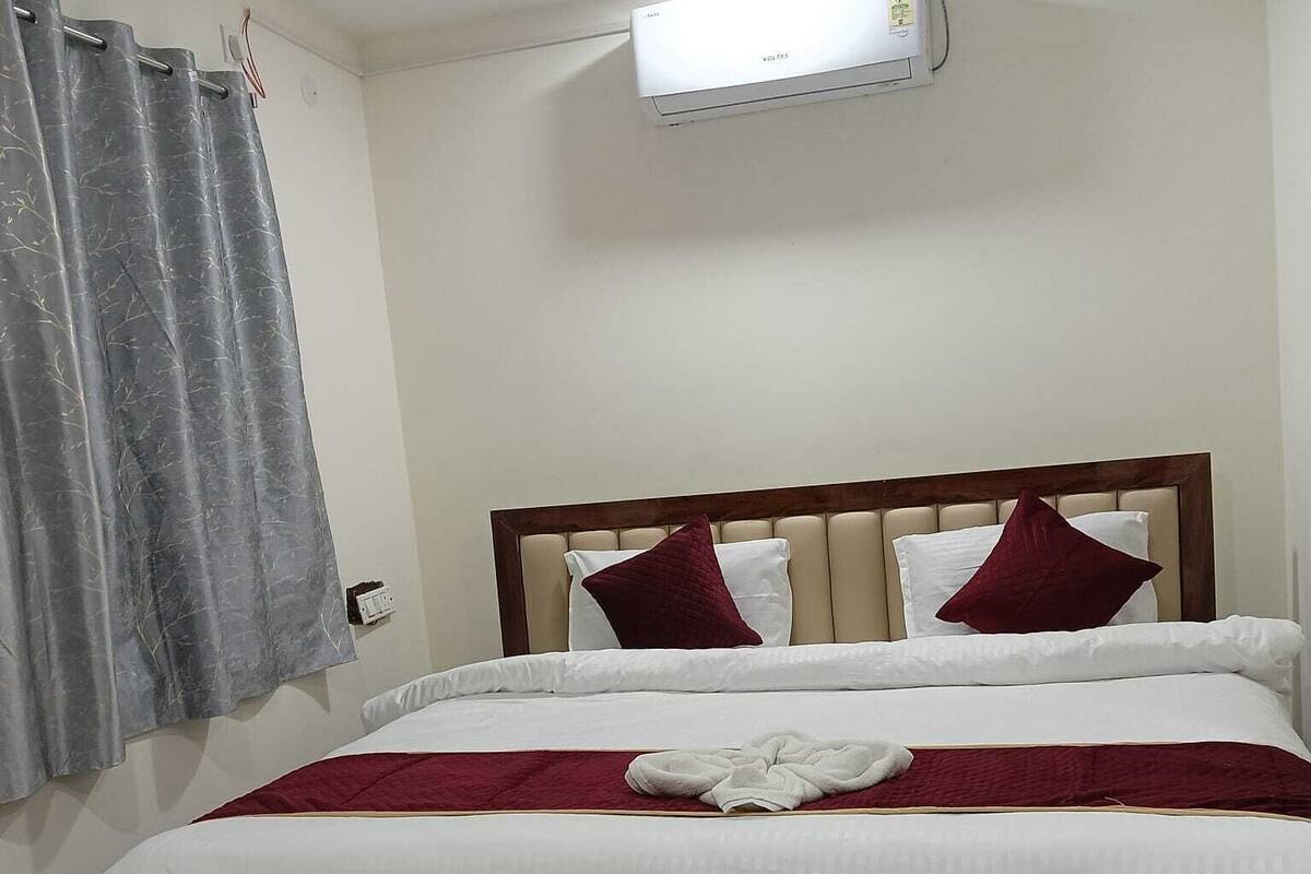 Una cama bien tendida con ropa blanca y almohadas rojas, una cortina gris y un aire acondicionado montado en la pared.