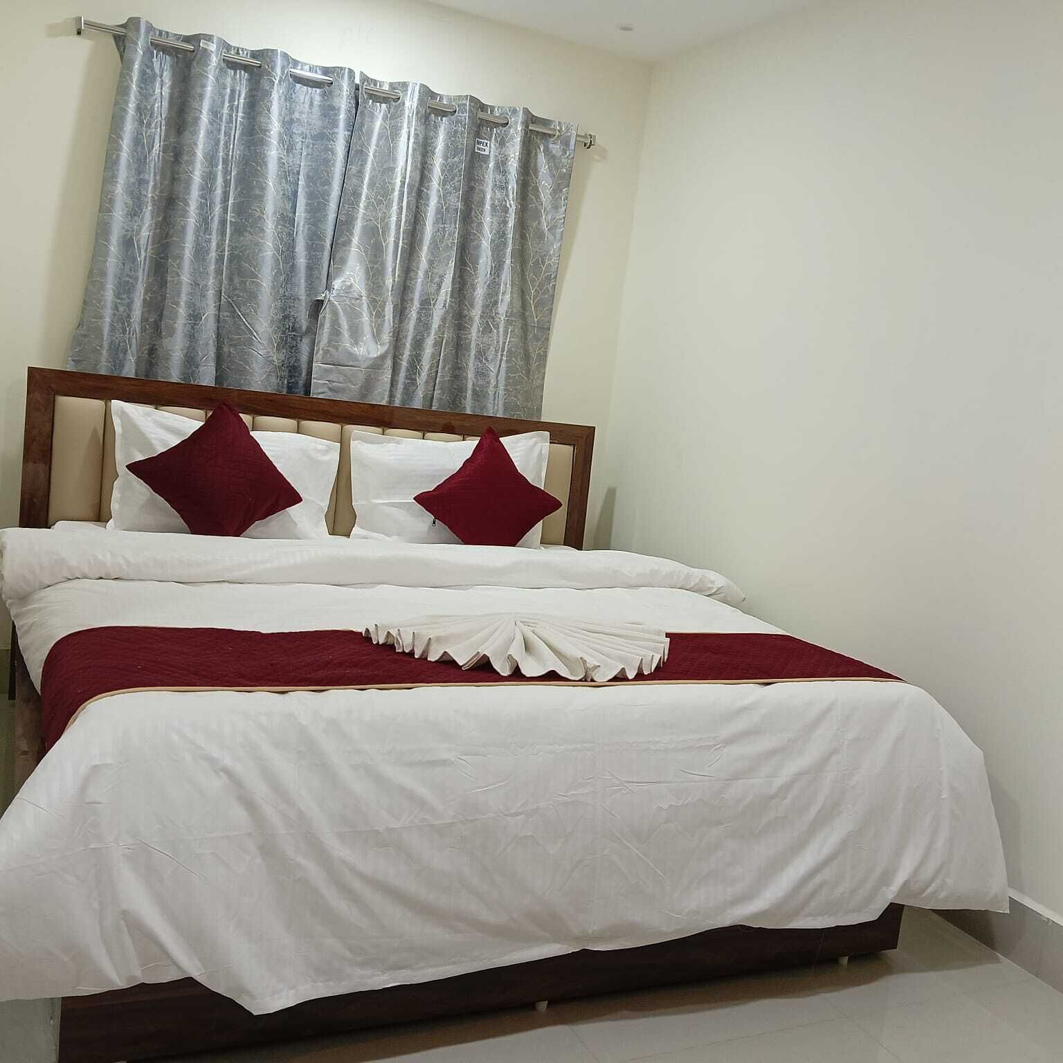 Una cama bien hecha con ropa de cama blanca y detalles rojos, un cabecero de madera y una cortina gris.
