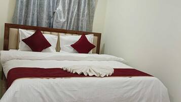 Una cama bien hecha con ropa de cama blanca y detalles rojos, un cabecero de madera y una cortina gris.