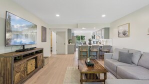 Een moderne woonkamer met een flatscreen televisie, houten meubels, een grijze bank en een keuken met een eettafel en stoelen.