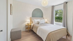 Een slaapkamer met een bed, twee nachtkastjes, een hanglamp, een raam met gordijnen en een klein voetenbankje.