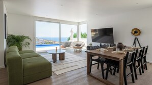 Una sala de estar moderna con un televisor grande, una mesa de comedor preparada para cuatro personas, un sofá verde y vistas al mar a través de amplias ventanas.