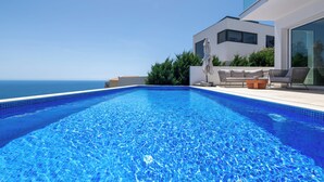 Una piscina moderna con agua azul cristalina, rodeada por un patio con mobiliario exterior y vistas al océano.