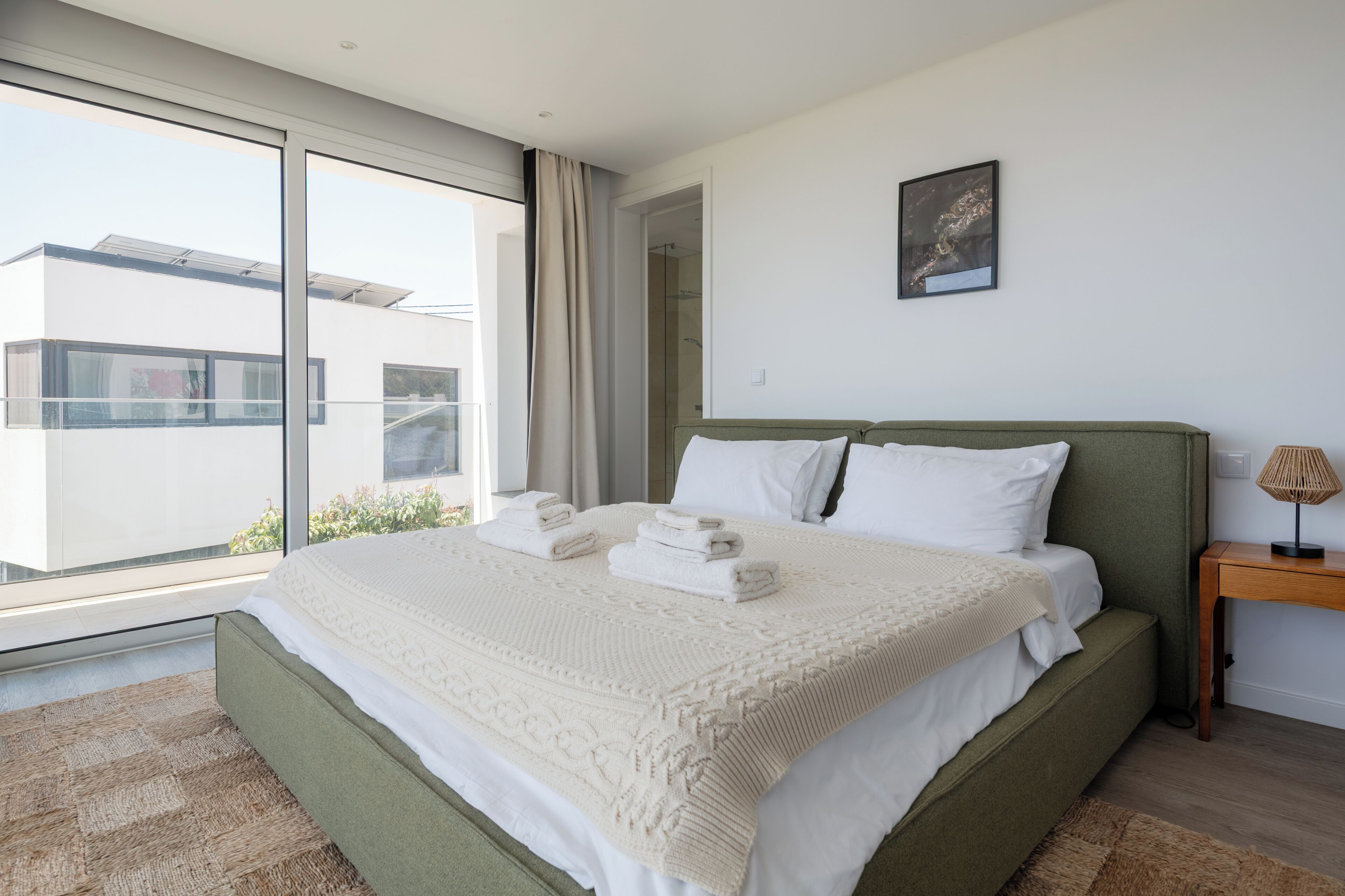 A modern bedroom with a large bed, a view of the outside, and a small bedside table with a lamp.