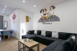 A modern living room with a black sectional sofa, a wooden coffee table, and a dining area with a table and chairs. - Palacio Real DownTown. 5BR 3BTH. NEW (Madrid)