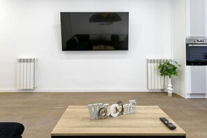 Ett modernt vardagsrum med en platt-TV som är monterad på väggen, ett träbord som fungerar som soffbord och som har en liten skylt med texten ”Välkommen”, en fjärrkontroll och en krukväxt.