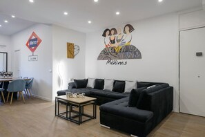 Ein modernes Wohnzimmer mit einem schwarzen Ecksofa, einem Couchtisch und einem Essbereich mit Holztisch und blauen Stühlen.