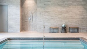 Un espace piscine intérieur moderne, avec un mur carrelé, une douche et deux petits bancs en bois.