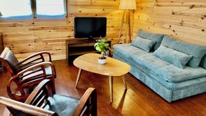 A cozy room with wooden walls, a sofa, a TV, a lamp, and a dining area with chairs and a table.