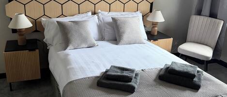 Een keurig opgemaakt bed met een geometrisch hoofdbord, twee nachtkastjes met lampen, een stoel en een groot abstract schilderij aan de muur.