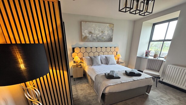 Une chambre d’hôtel moderne avec un grand lit, une tête de lit ornée d’un motif géométrique, une table de chevet avec une lampe, une chaise et une fenêtre donnant sur un paysage verdoyant.
