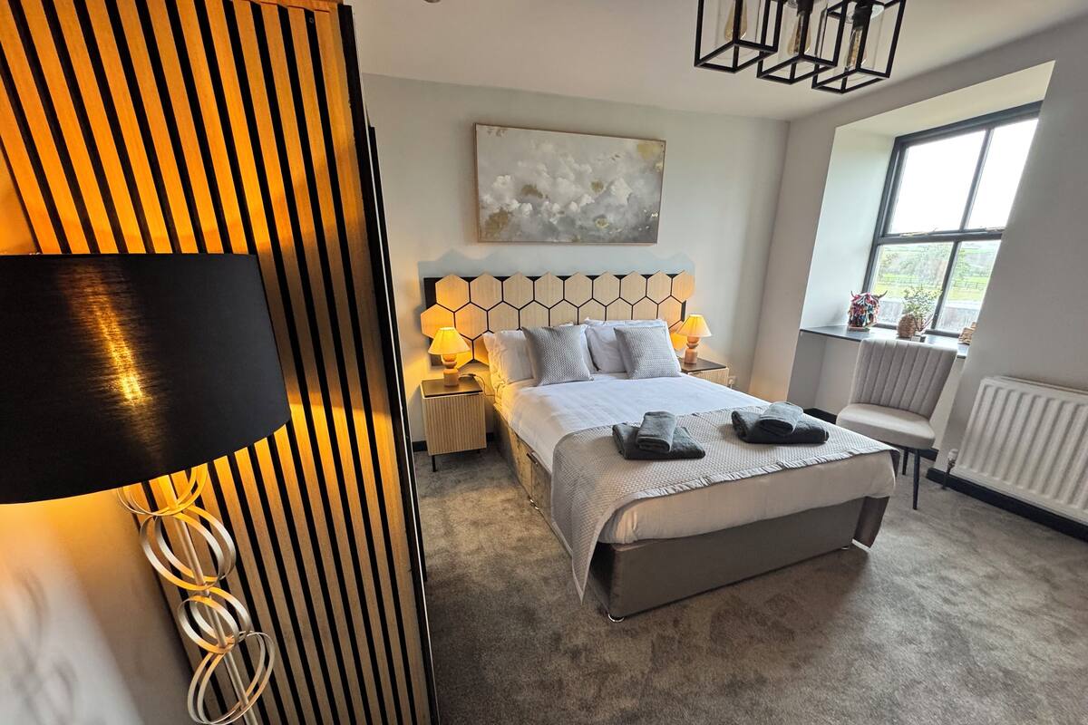 A modern hotel room with a large bed, a headboard with a geometric pattern, a bedside table with a lamp, a chair, and a window with a view of greenery.