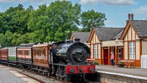 Une locomotive à vapeur d’époque se trouve dans une gare, avec un quai et un bâtiment doté d’une porte rouge.