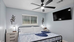A bedroom with a bed, bedside tables, a ceiling fan, a TV, and a window with blinds.