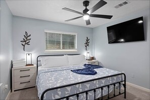 Une chambre à coucher comprenant un lit, des tables de chevet, un ventilateur de plafond, une télévision et une fenêtre avec des stores.