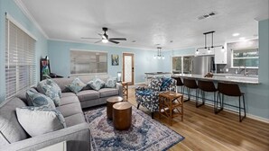 A living room with a gray sofa, patterned armchair, and round coffee table. There is a kitchen area with a bar and stools.