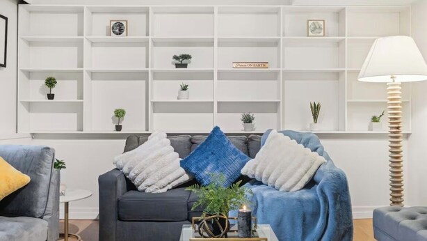 Uma sala de estar moderna com um sofá cinzento, uma poltrona azul e uma mesa de centro com um objeto decorativo. Estantes ao longo da parede exibem diversos objetos e plantas.