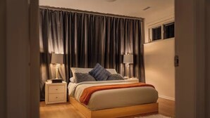 Quarto com cama de madeira, duas mesas de cabeceira com luminárias, tapete branco e cortinas escuras.