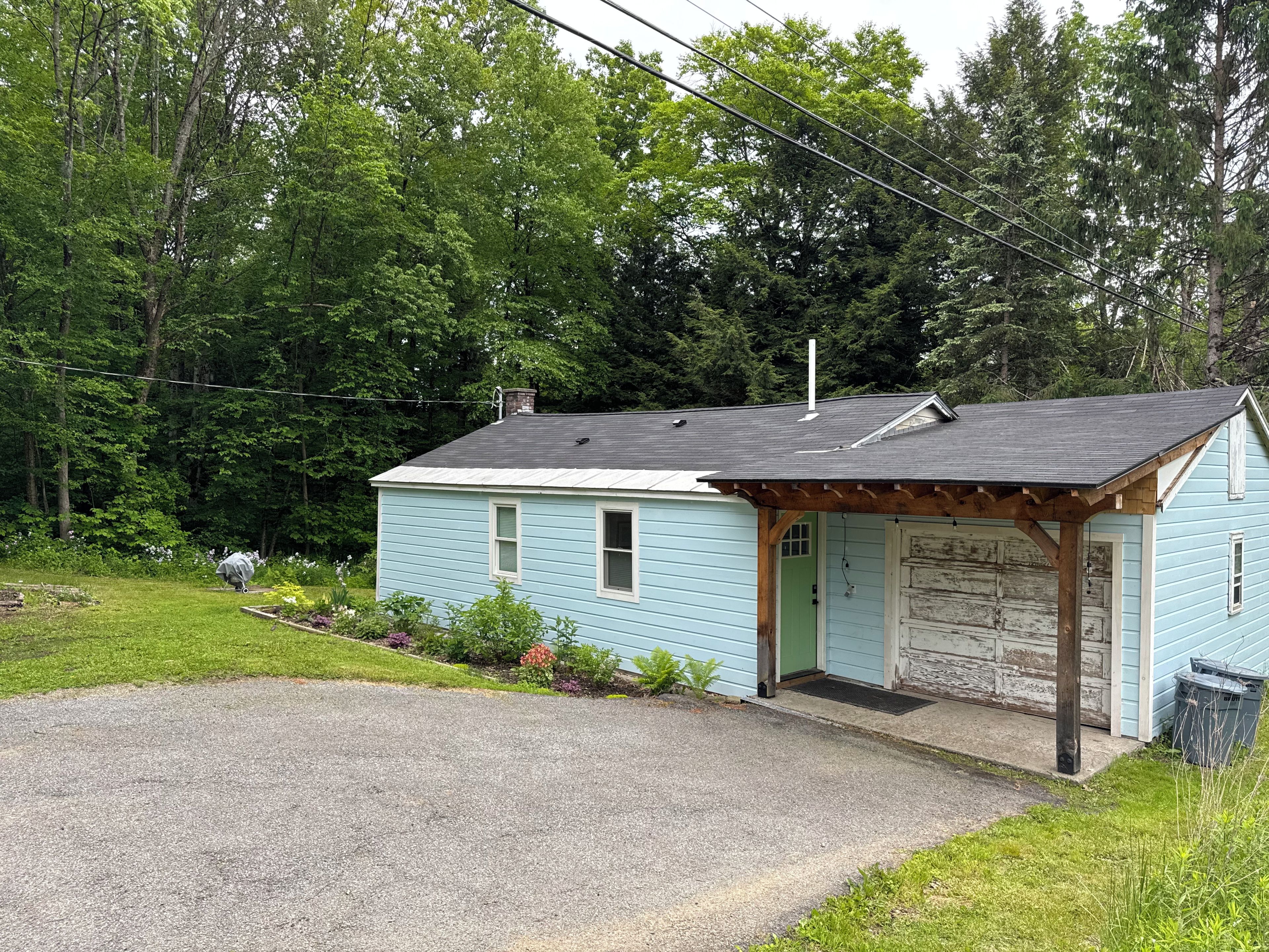 A small, light blue house with a porch and a garage, surrounded by a grassy yard and dense trees.