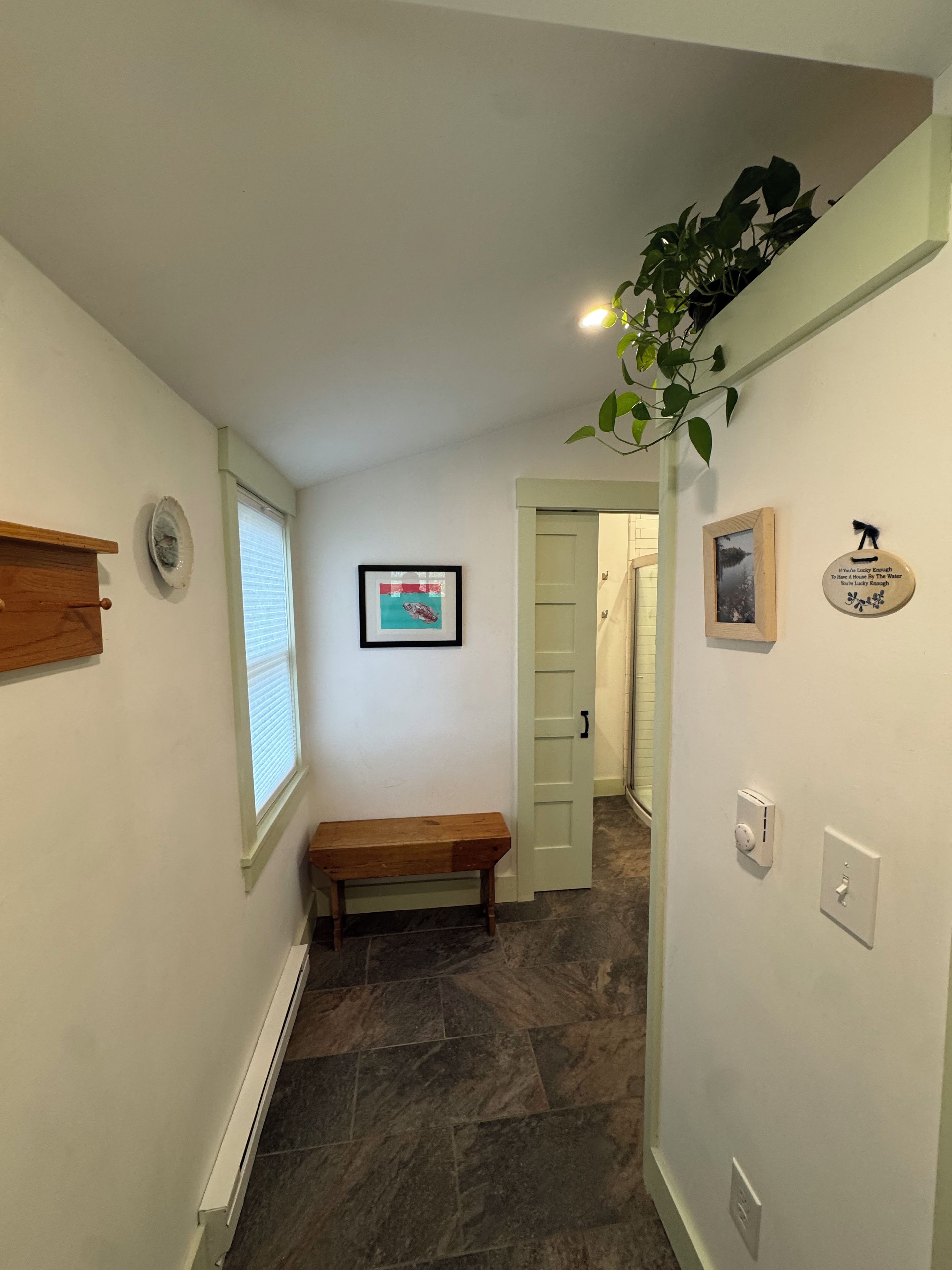 Un couloir avec un banc, un tableau encadré, une plante en pot et une porte donnant sur une autre pièce.