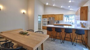 A modern kitchen with wooden cabinets, a central island with bar stools, and a dining area with a wooden table and chairs. - Parkview Place - Wine Country Home with Jetted Tub (McMinnville)