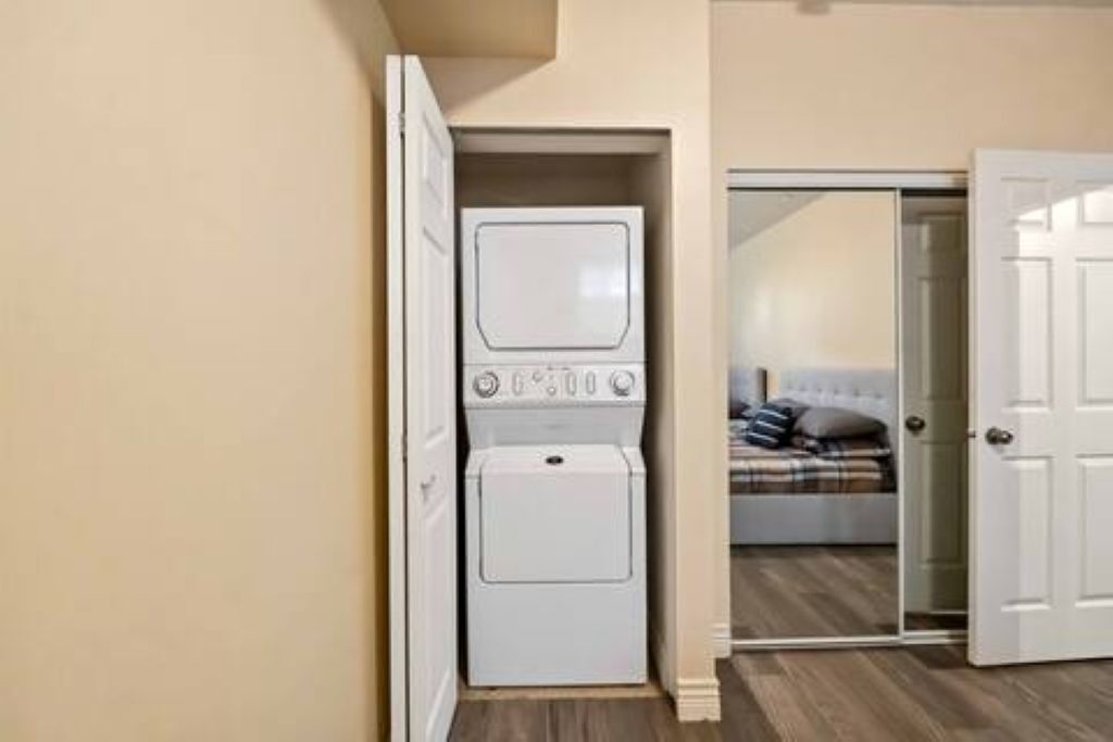 A laundry room with stacked washers and dryers, a bedroom with a bed and a nightstand, and a door leading to another room.