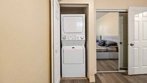 Una lavandería con lavadoras y secadoras apiladas, un dormitorio con cama y mesita de noche, y una puerta que da a otra habitación.