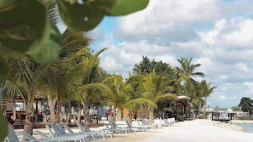 A beach with lounge chairs and palm trees, a wooden walkway, and a building in the background.