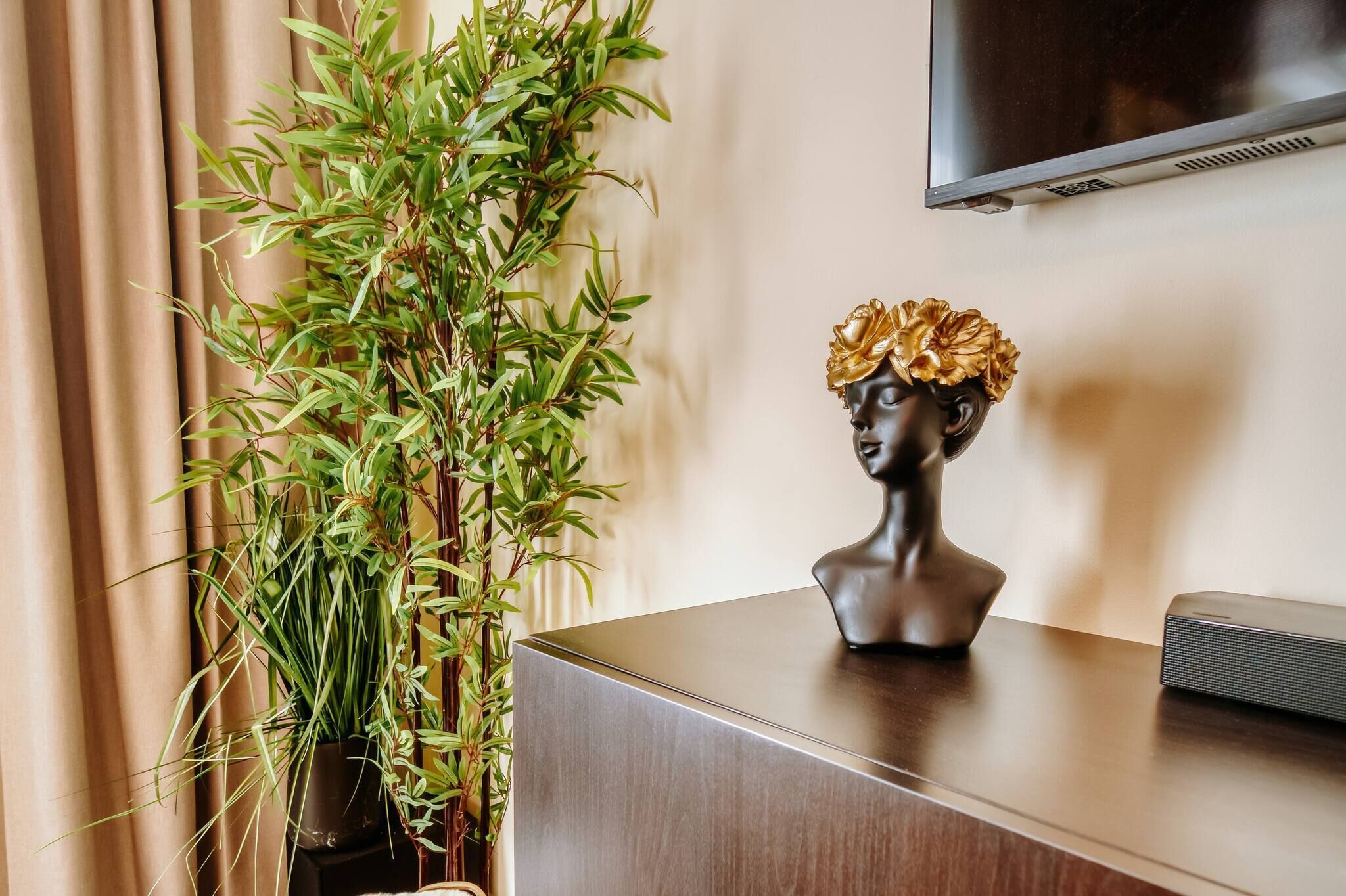 Una habitación moderna con un escritorio de madera, una planta en maceta, una escultura de busto y un televisor de pantalla plana montado en la pared.