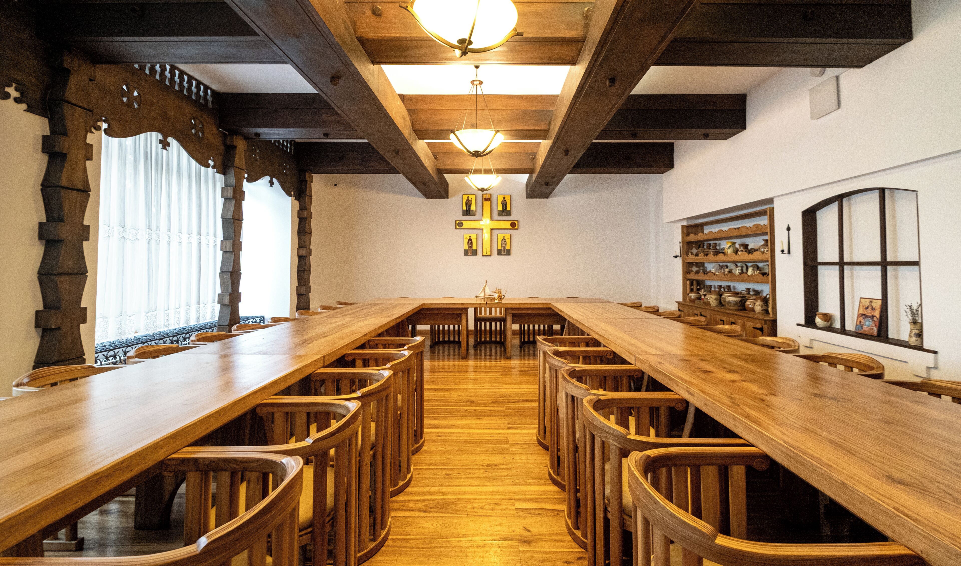 A long wooden table with chairs, a cross on the wall, and a window with shelves.
